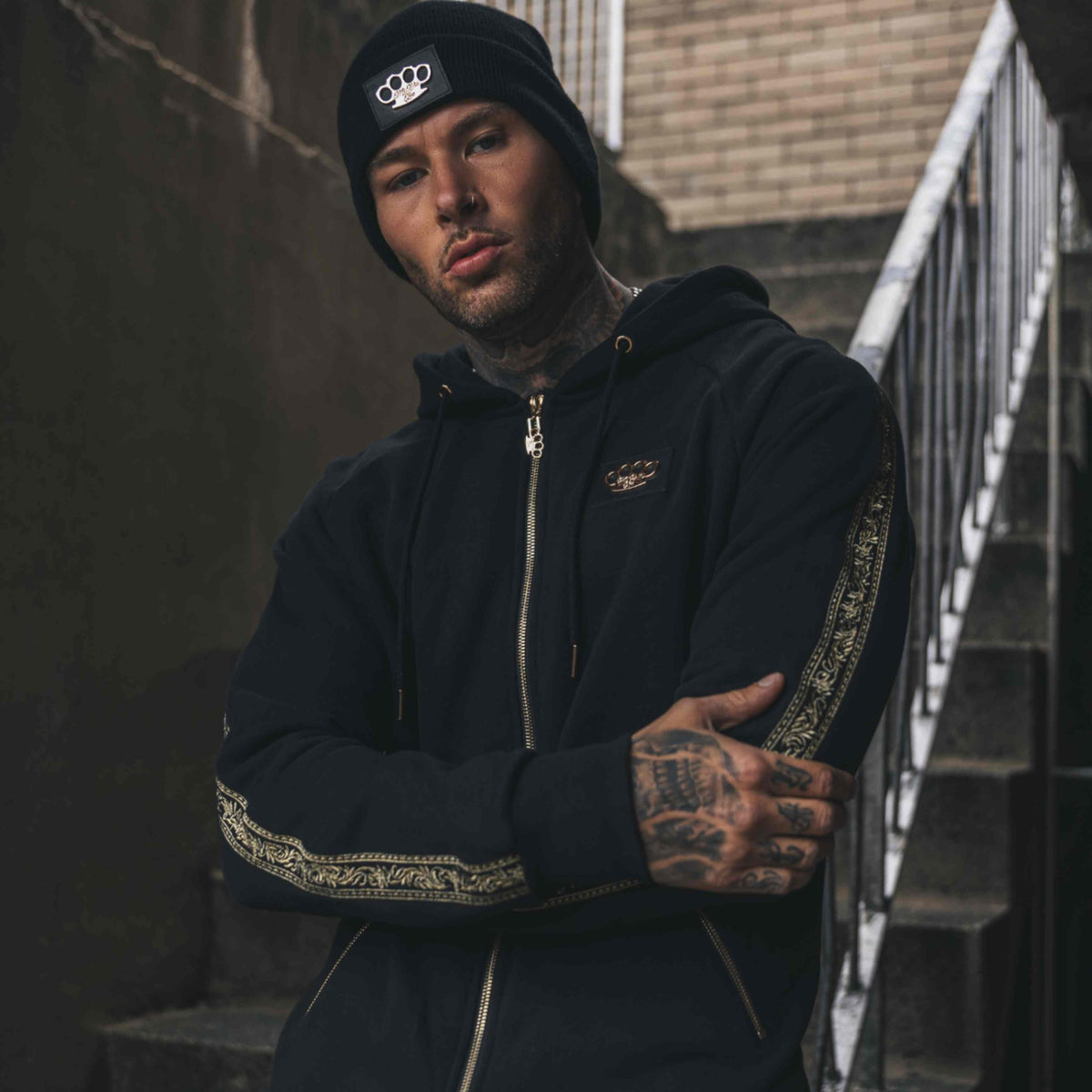 Model in zwart MVL Meander trainingspak met gouden embroidery band op de mouwen, op een trap
