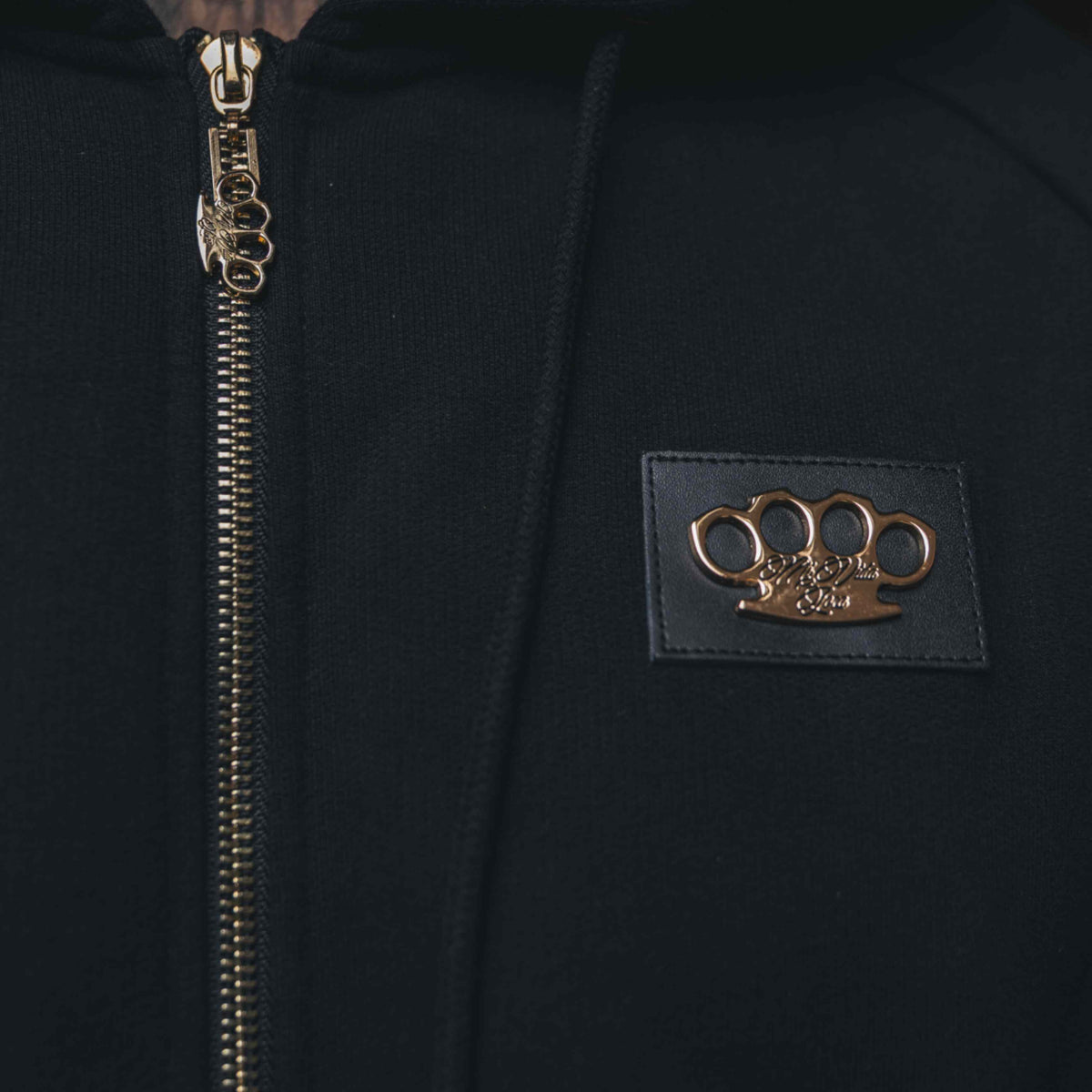 Close-up van het gouden MVL logo op lederen patch en de gouden rits van de Meander hoodie