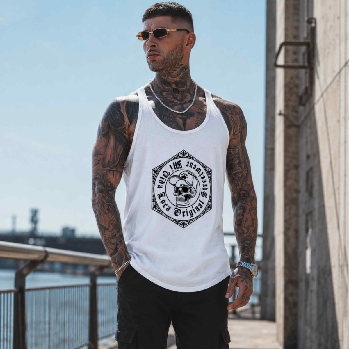 Model met tattoos en zonnebril draagt de witte MVL Original streetwear tanktop aan het water